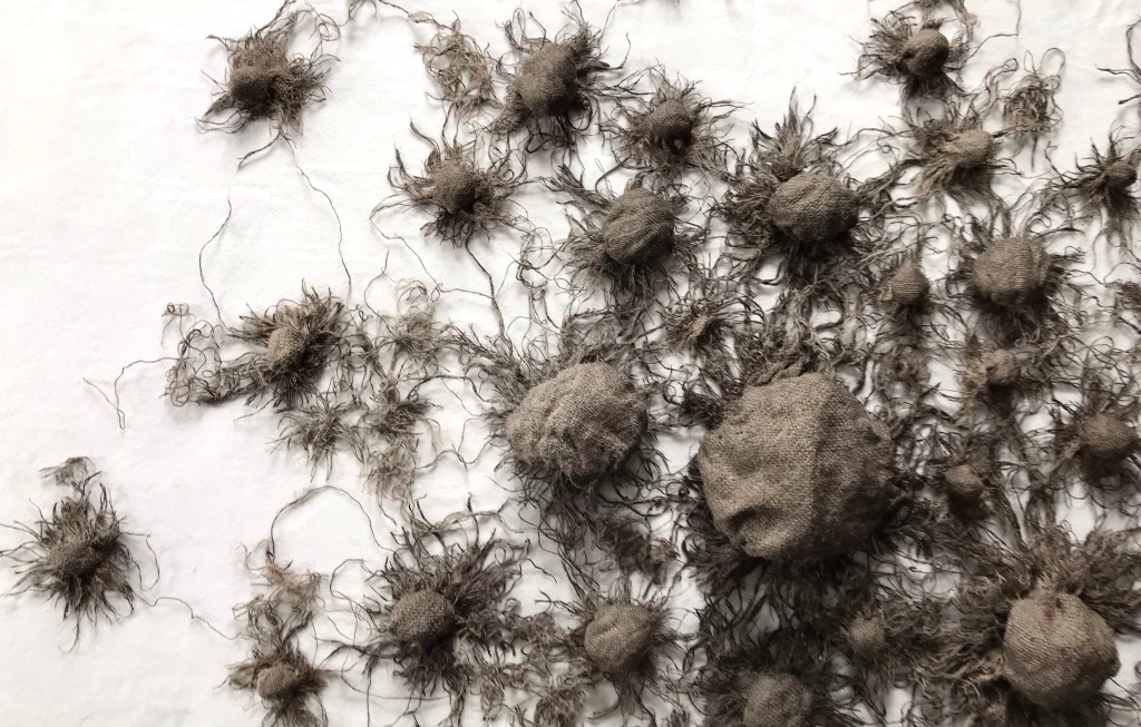 Detail of a large art installation piece by MJ Seal made from sackcloth stained with charred bones that resembles a giant cluster of dark gray puffball mushrooms with extending roots emerging from a wall